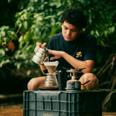 A young man carefully brews coffee using a pour-over method near a lush forest stream.